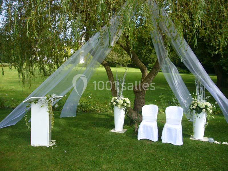 Décoration de cérémonie en plein air avec chaises blanches, urnes fleuries et voilage suspendu sous un arbre.