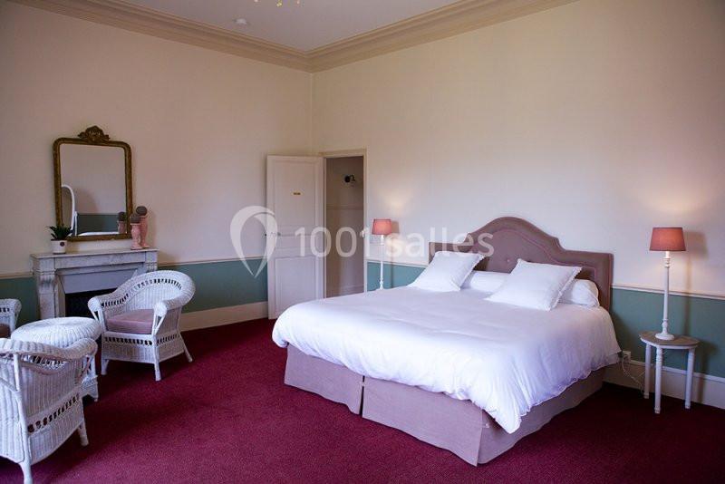 Chambre lumineuse avec lit double, fauteuils en osier, cheminée, miroir et lampes sur un tapis rouge.