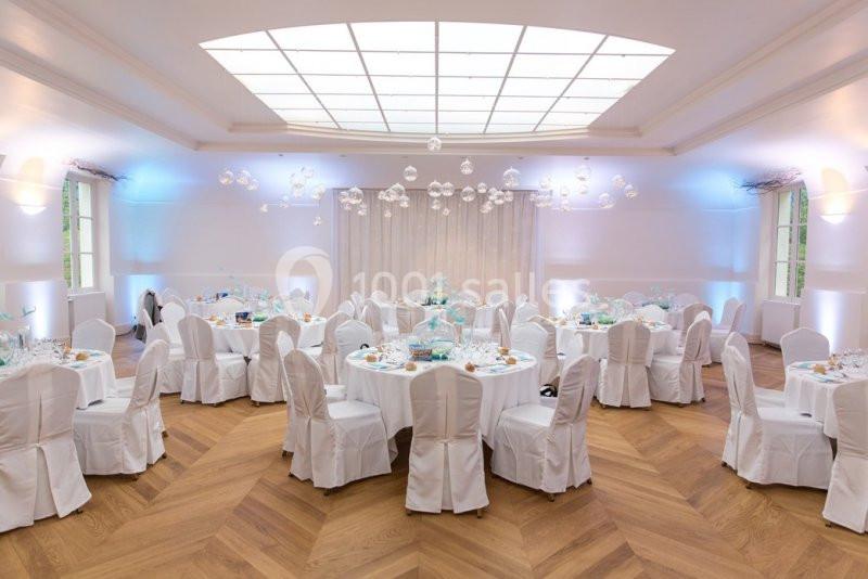 Salle de réception lumineuse avec tables rondes dressées, chaises blanches et décorations suspendues.