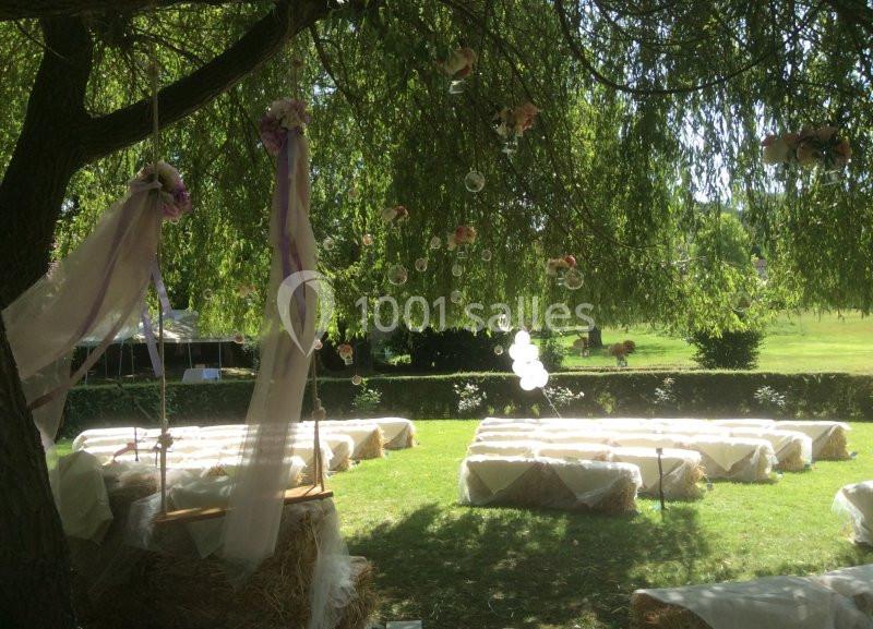 Chaises en bottes de paille décorées sous un arbre pour une cérémonie en extérieur, avec guirlandes et ballons blancs.