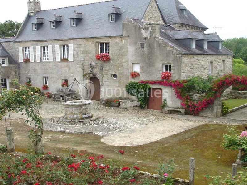 Maison en pierre traditionnelle avec des volets blancs, un puits central et des fleurs rouges sur la façade.