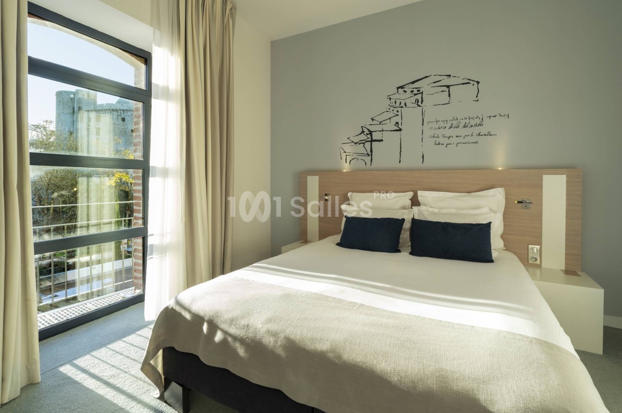Chambre lumineuse avec un lit double, deux coussins bleus, une grande fenêtre et une décoration murale minimaliste.