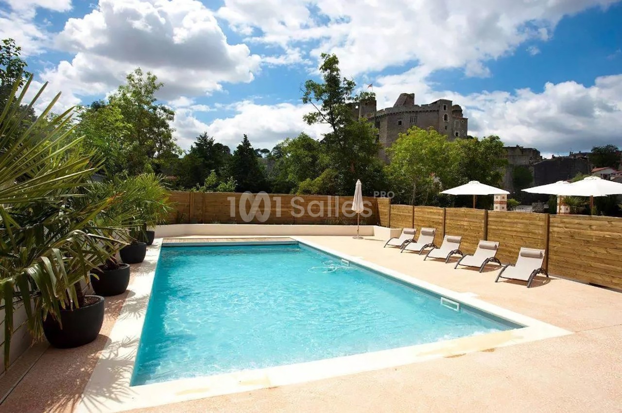 Piscine extérieure entourée de transats, parasols, palmiers et clôture en bois, avec un château en arrière-plan.