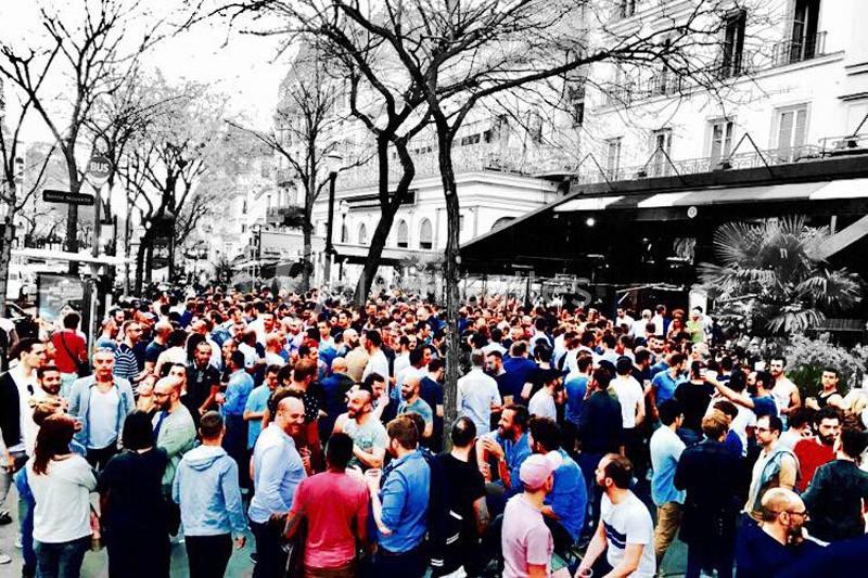 Foule rassemblée à l'extérieur d'un bâtiment, dans une rue bordée d'arbres et d'immeubles.