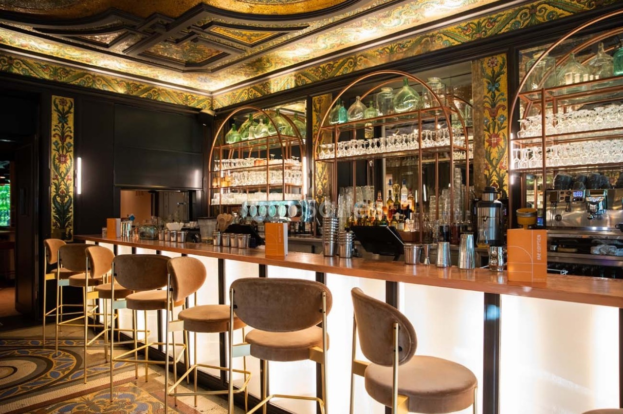 Bar élégant avec comptoir lumineux, tabourets rembourrés, étagères garnies de verres et décorations murales ornées.