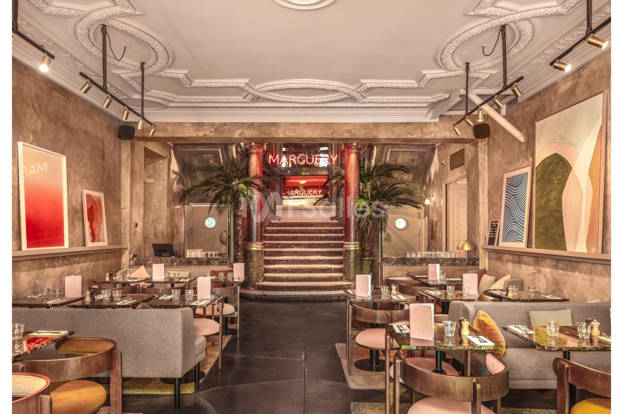Salle de restaurant élégante avec des tables dressées, un escalier central, des plantes et des œuvres d'art aux murs.