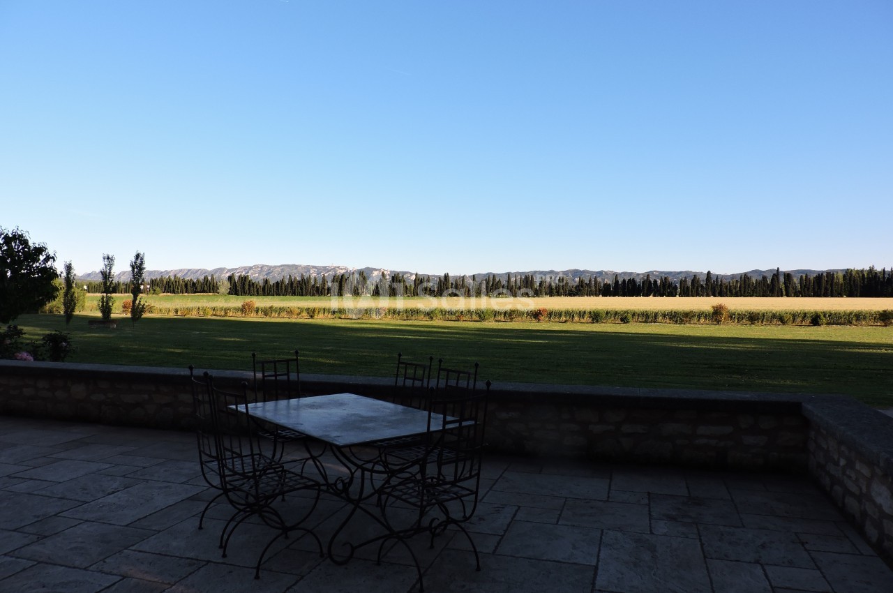 Vue sur un champ ouvert bordé d'arbres, depuis une terrasse en pierre avec une table et des chaises en métal.