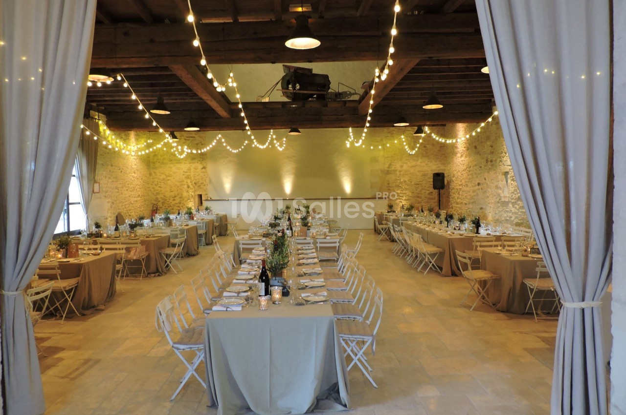 Grande salle de réception décorée avec des guirlandes lumineuses, des tables dressées et des chaises pliantes.