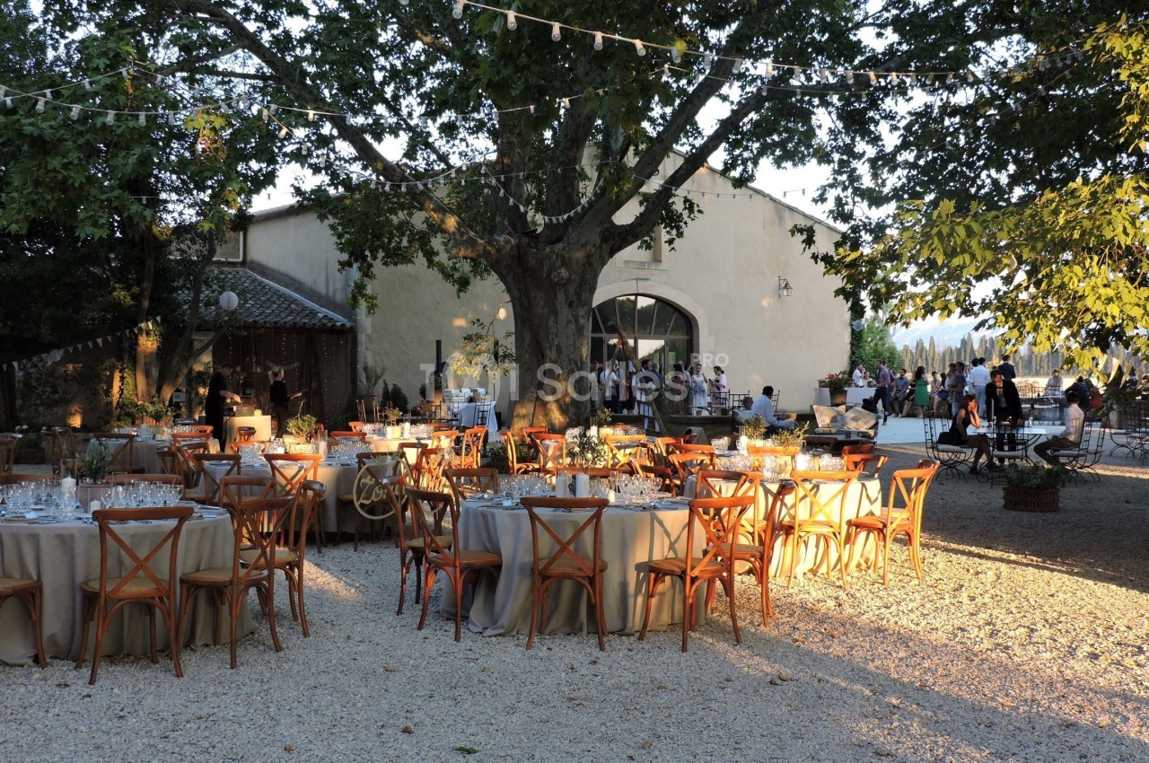 Tables dressées pour un événement en plein air sous des arbres, avec des guirlandes lumineuses et un bâtiment en arrière…