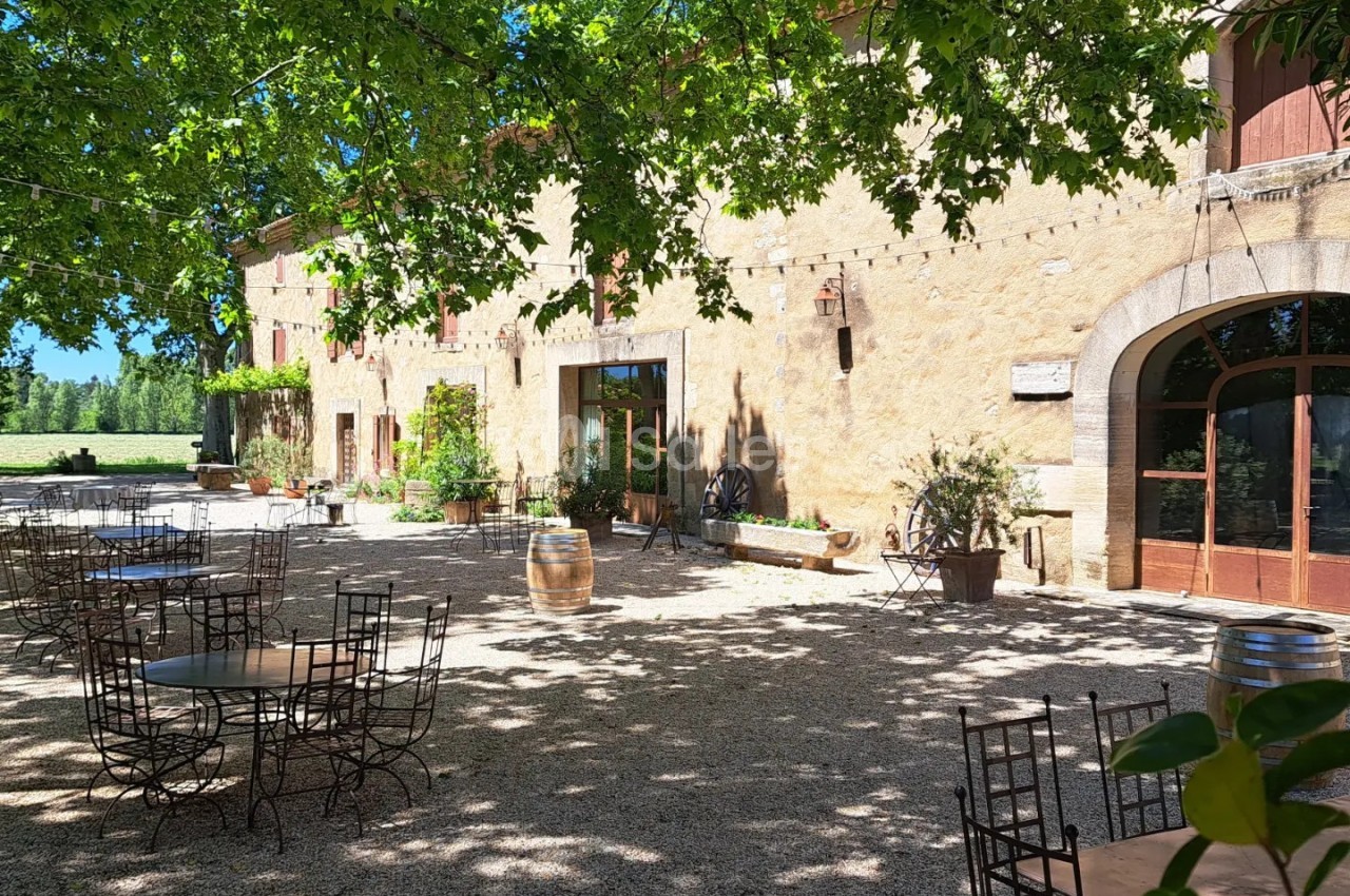 Cour ombragée d'une bâtisse en pierre avec tables et chaises en métal, entourée de végétation et baignée de lumière…