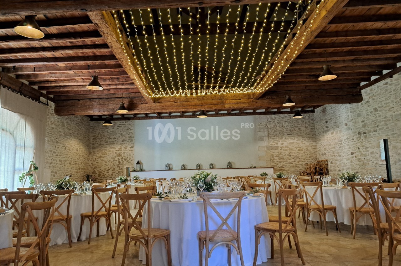 Salle de réception avec tables rondes dressées, chaises en bois et plafond décoré de guirlandes lumineuses.