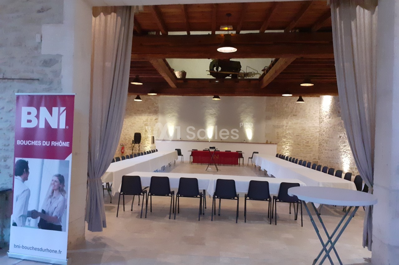 Salle de réunion avec tables disposées en U, chaises noires, murs en pierre, plafond en bois et un kakemono ’BNI’.
