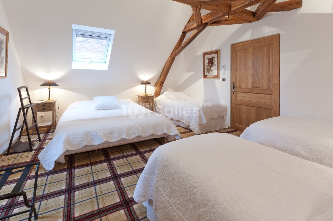 Chambre mansardée avec trois lits simples, décorée de poutres apparentes, éclairée par une fenêtre de toit.