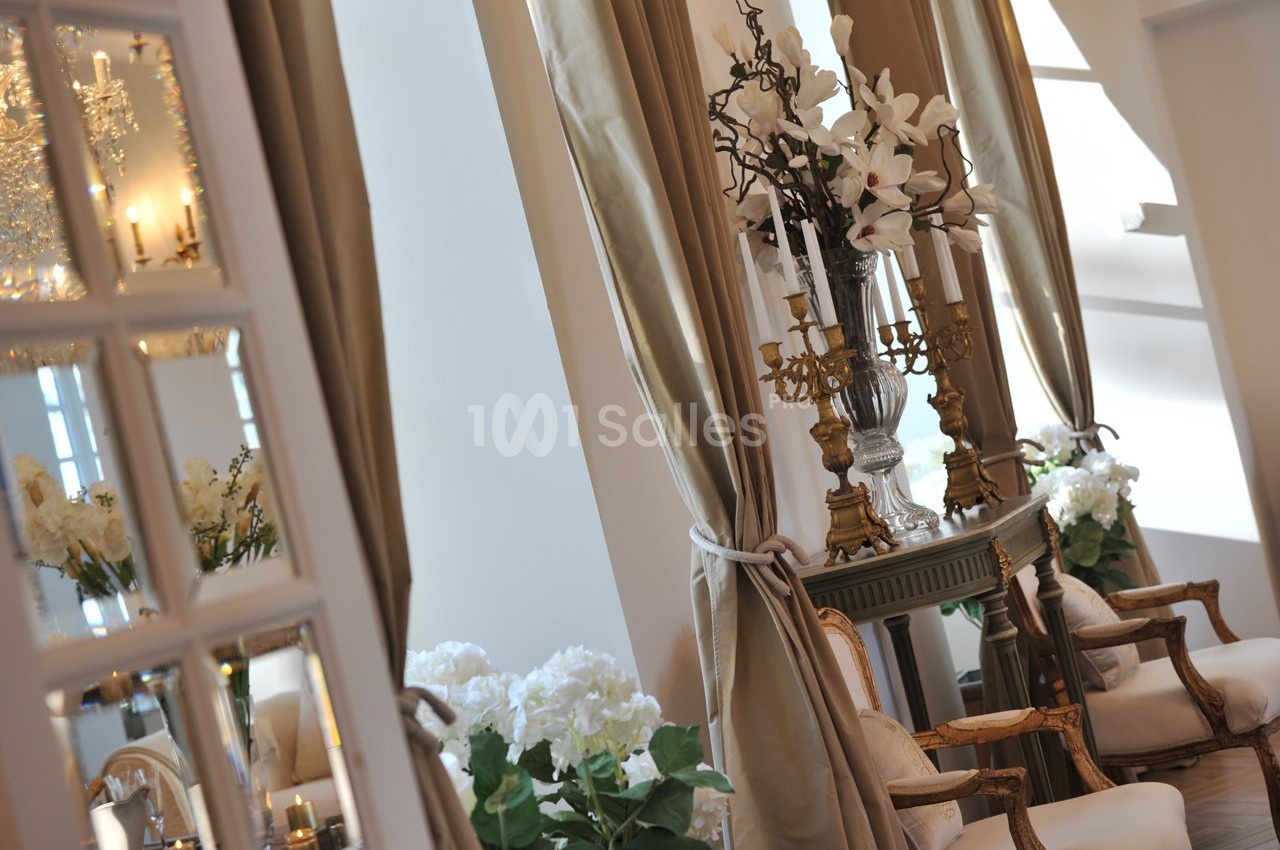 Salle élégante avec fauteuils classiques, rideaux beiges, fleurs blanches et chandelier doré sur une console.