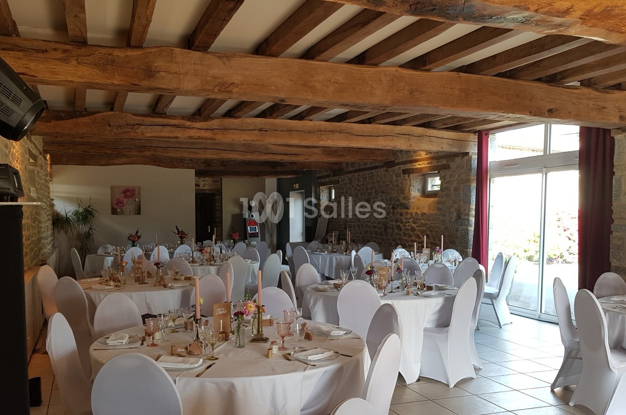 Salle de réception avec des tables rondes dressées, chaises blanches et poutres apparentes au plafond.