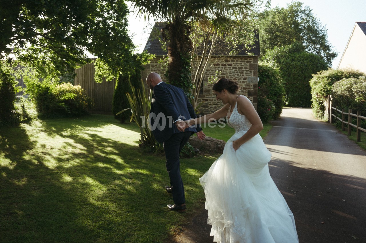 Un couple en tenue de mariage marche sur une allée bordée de verdure, éclairée par une lumière naturelle.