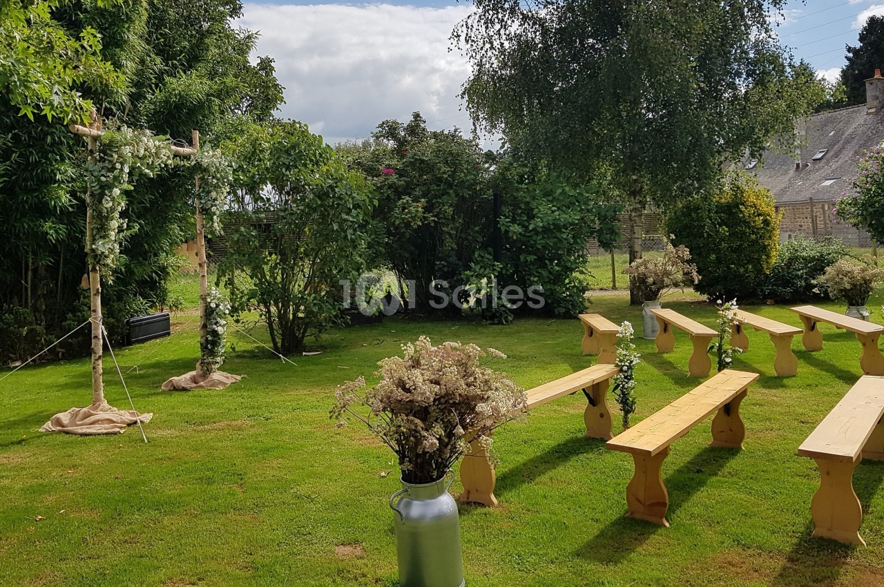Chaises en bois disposées en extérieur devant une arche fleurie, dans un jardin verdoyant sous un ciel partiellement nuageux.