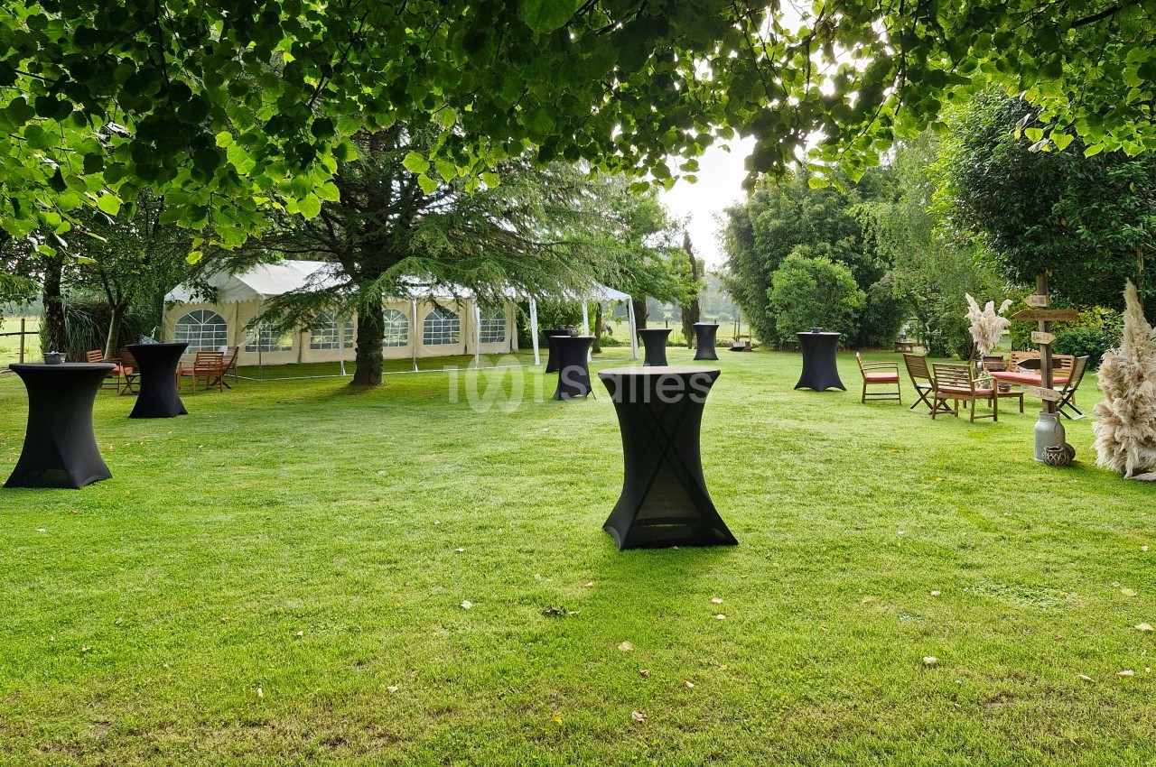 Jardin avec tables hautes noires, chaises en bois et une tente blanche en arrière-plan, entouré de verdure.