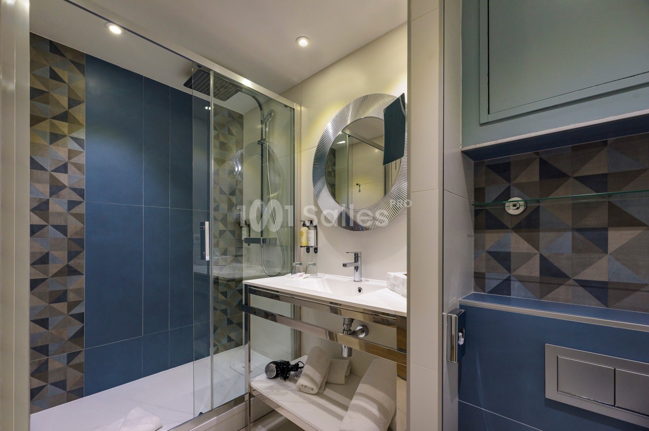 Salle de bain moderne avec douche à l'italienne, carrelage géométrique, miroir rond et lavabo intégré.