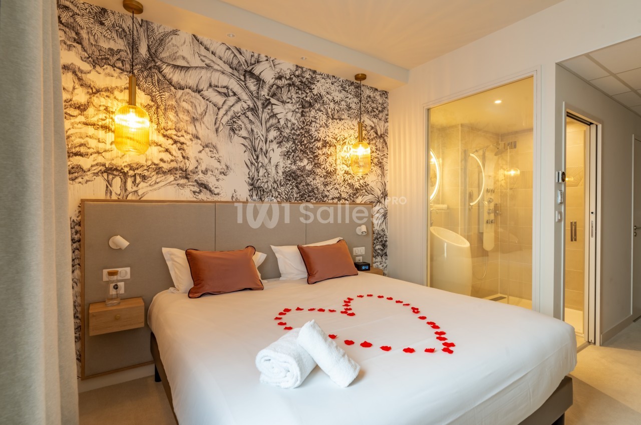 Chambre d'hôtel avec lit double décoré de pétales rouges en forme de cœur, éclairage chaleureux et salle de bain attenante.