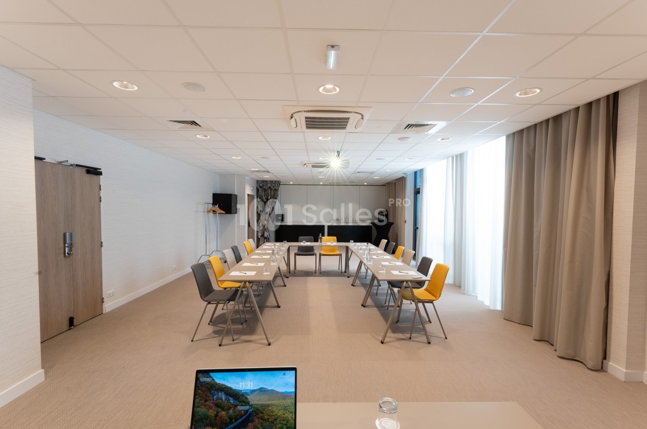 Salle de réunion lumineuse avec tables disposées en U, chaises colorées et équipement audiovisuel.