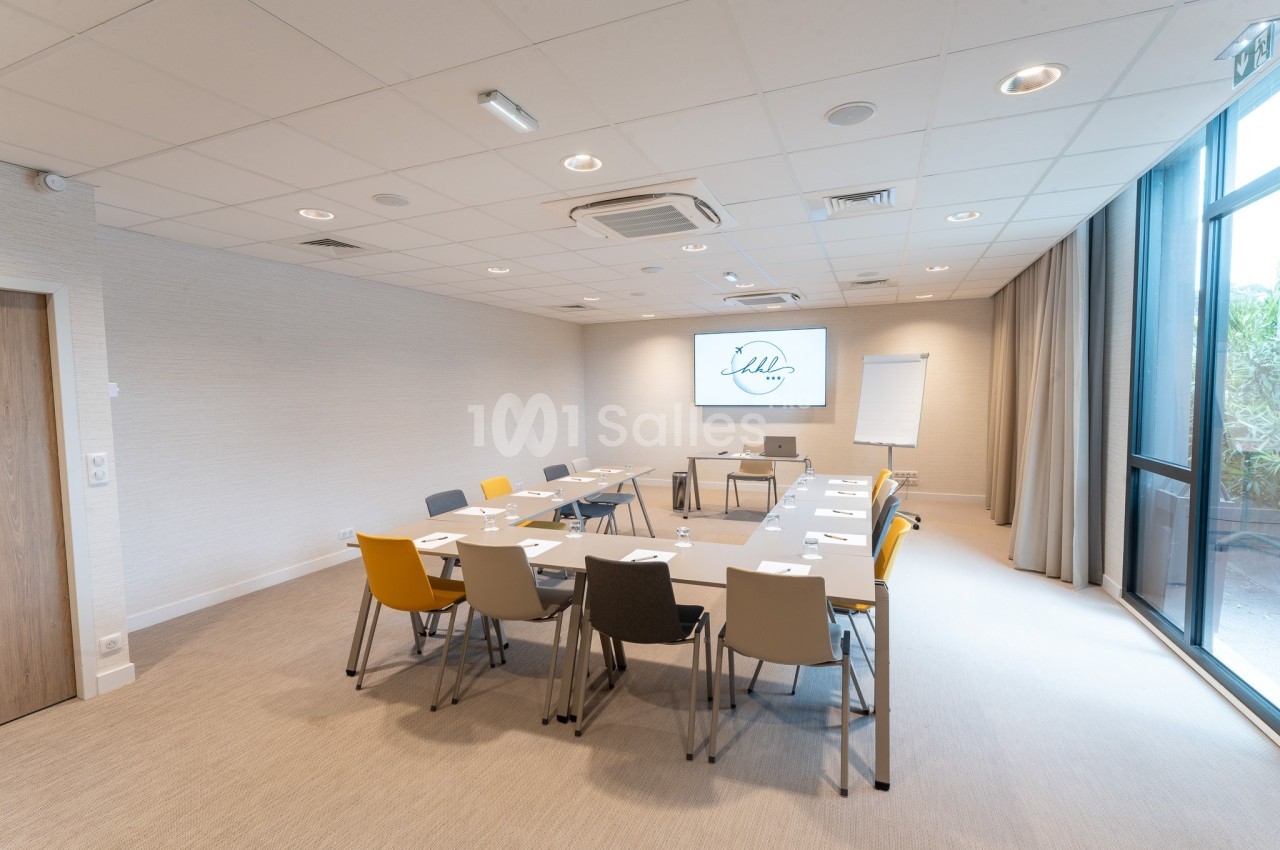 Salle de réunion lumineuse avec table rectangulaire, chaises colorées, écran, paperboard et grande baie vitrée.