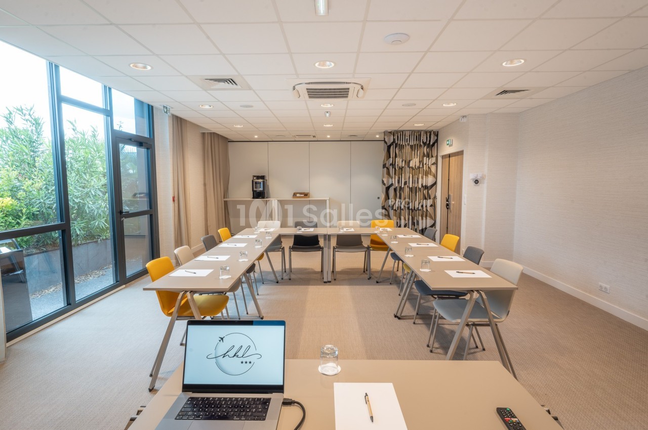 Salle de réunion lumineuse avec tables disposées en U, chaises colorées, écran d'ordinateur et machine à café en arrière…