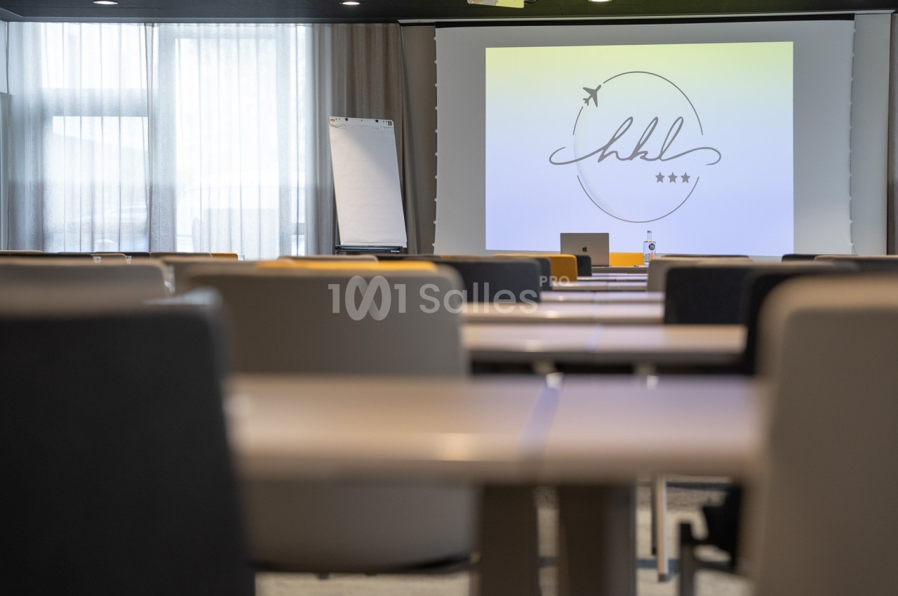 Salle de conférence lumineuse avec des rangées de tables, un écran de projection et un tableau blanc en arrière-plan.