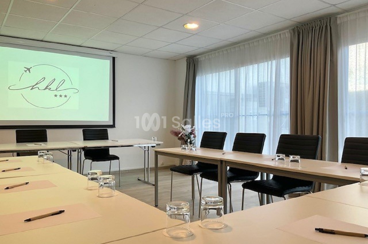 Salle de réunion lumineuse avec tables disposées en U, écran de projection allumé et chaises noires.