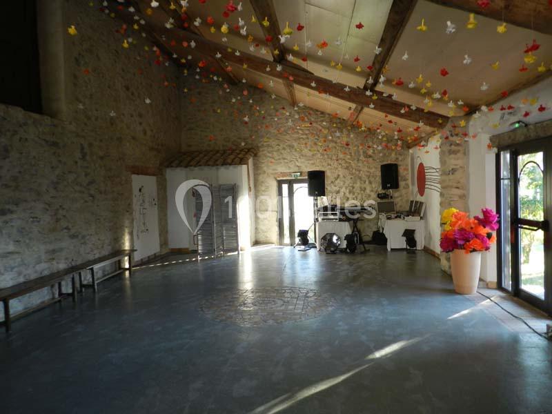 Salle en pierre avec plafond en bois, décorée de guirlandes colorées suspendues, équipée d'un coin sono et d'un vase de…