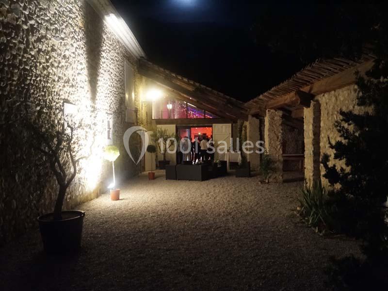 Cour intérieure en soirée avec des murs en pierre éclairés, menant à une salle illuminée où des personnes sont réunies.