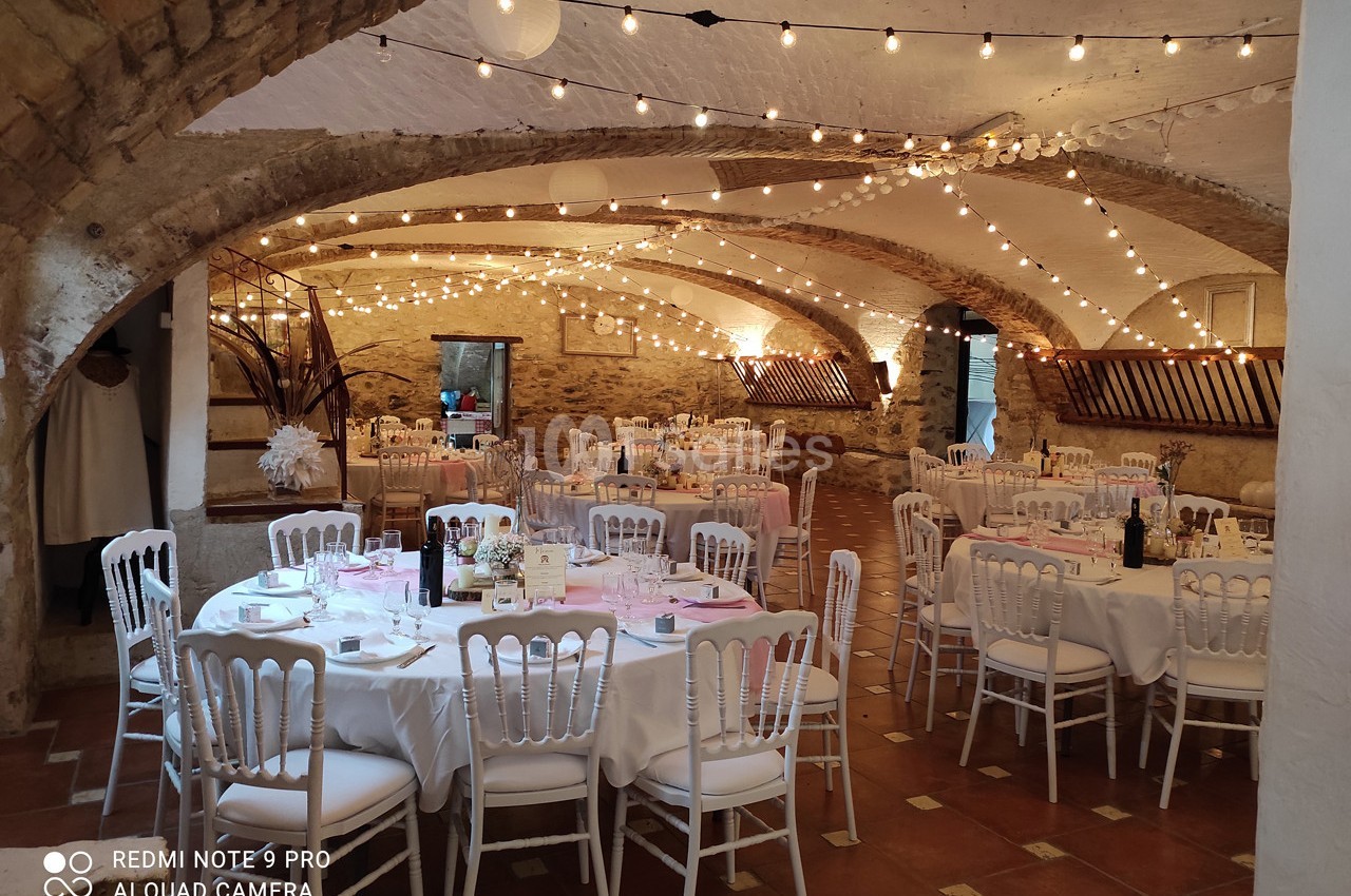Salle voûtée décorée avec des guirlandes lumineuses, tables rondes dressées pour un événement festif.
