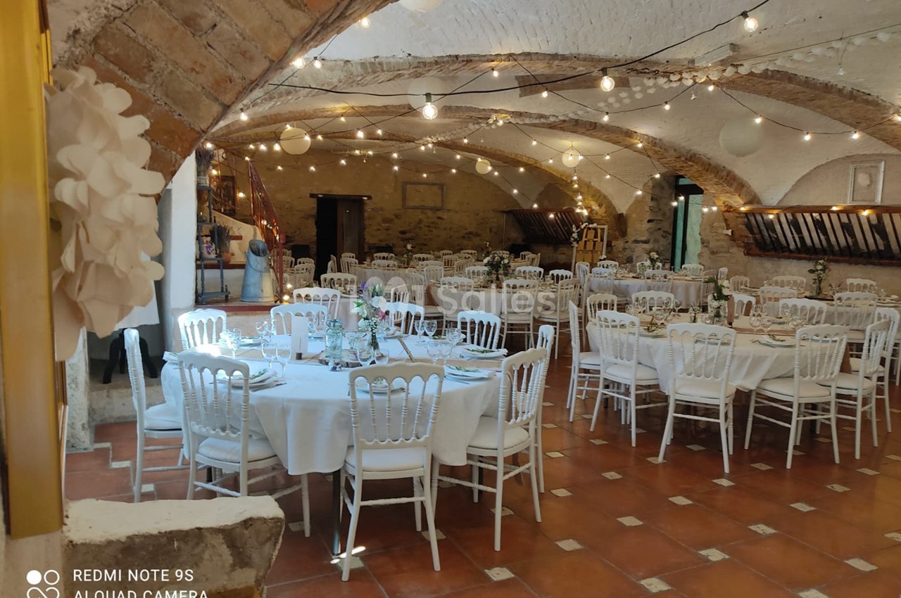 Salle voûtée décorée pour un événement, avec tables rondes dressées, chaises blanches et guirlandes lumineuses suspendues.