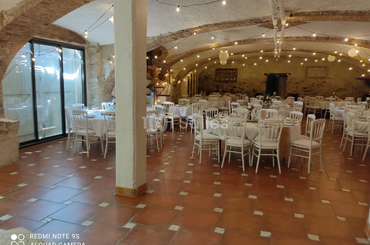 Salle de réception avec tables rondes dressées, chaises blanches et guirlandes lumineuses sous un plafond voûté.