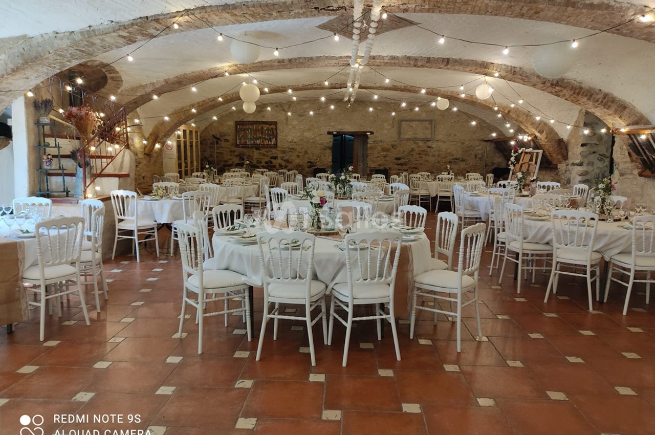 Salle voûtée décorée pour un événement, avec des tables rondes dressées et des guirlandes lumineuses suspendues.