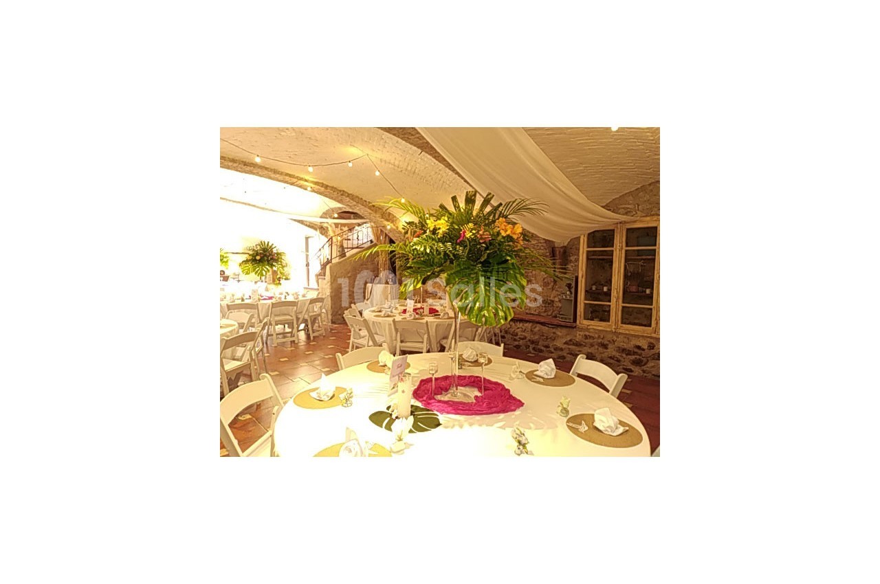 Salle décorée pour un événement avec des tables rondes, nappes blanches, centres de table fleuris et éclairage chaleureux.