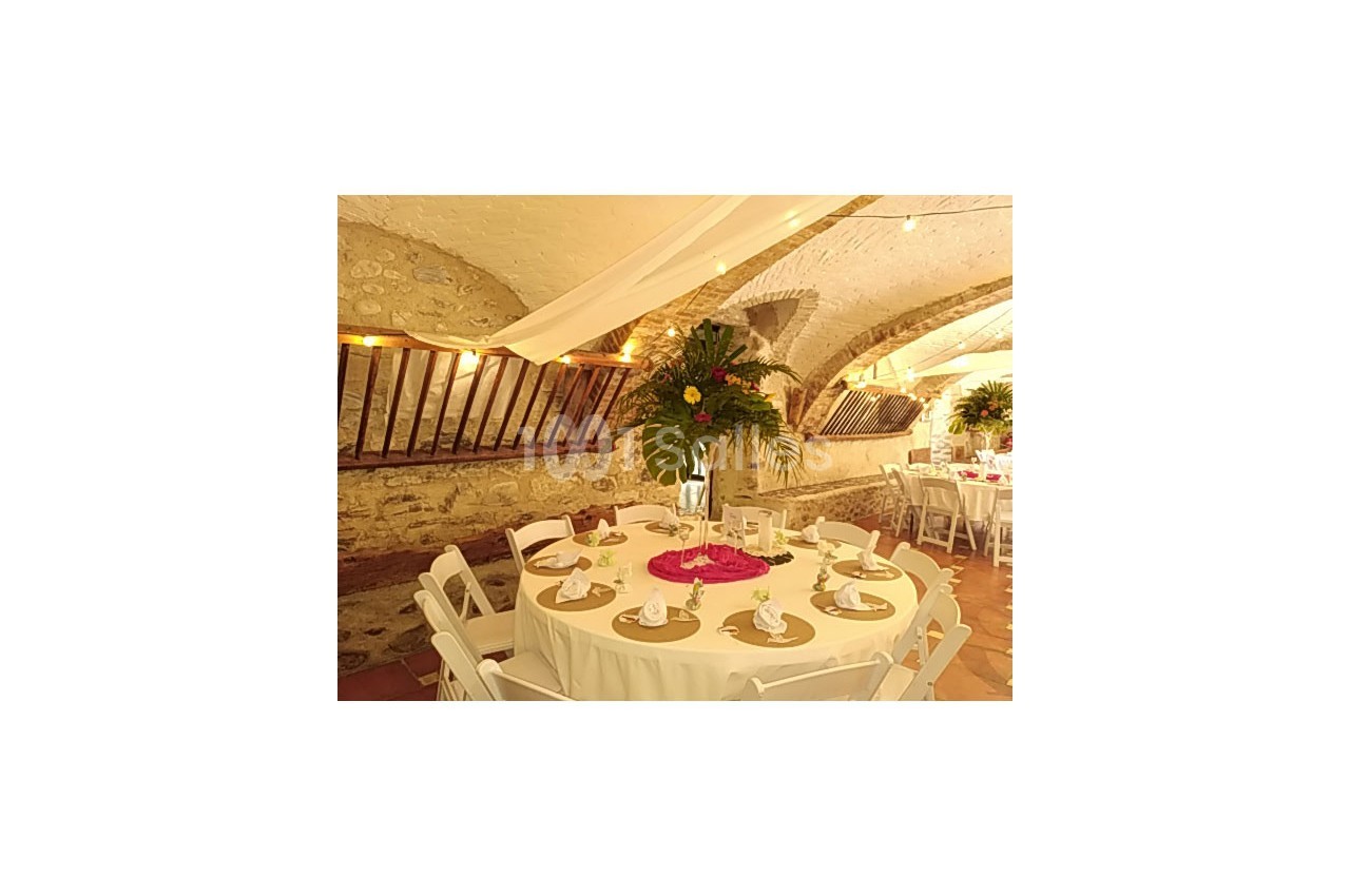 Salle voûtée décorée pour un événement, avec tables rondes dressées, nappes blanches et centres de table fleuris.