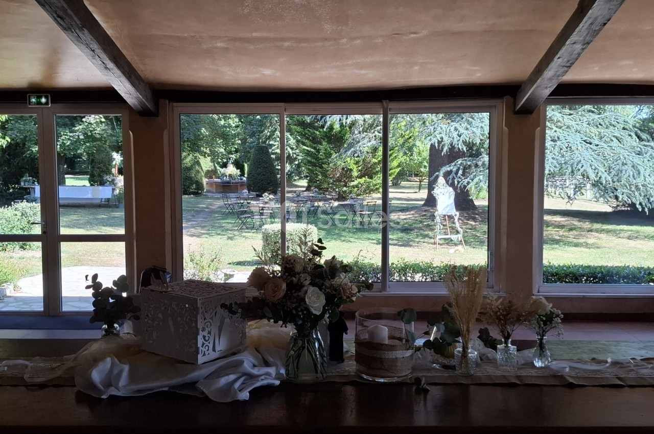 Vue intérieure d'une salle avec une table décorée de fleurs, donnant sur un jardin verdoyant à travers de grandes baies…