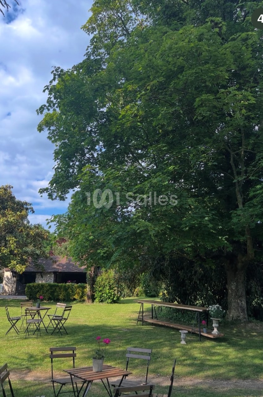 Tables et chaises en métal disposées sur une pelouse verdoyante, sous un grand arbre, par une journée ensoleillée.