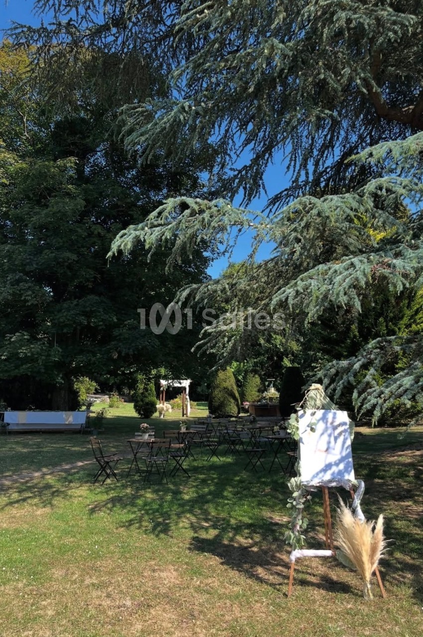 Chevalet décoré et chaises disposées dans un jardin ombragé, entouré d'arbres et de verdure.