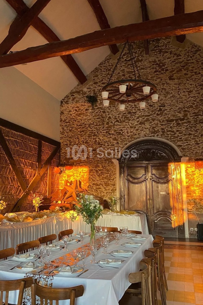 Salle de réception rustique avec murs en pierre, poutres apparentes, tables dressées et lumière chaude du coucher de soleil.