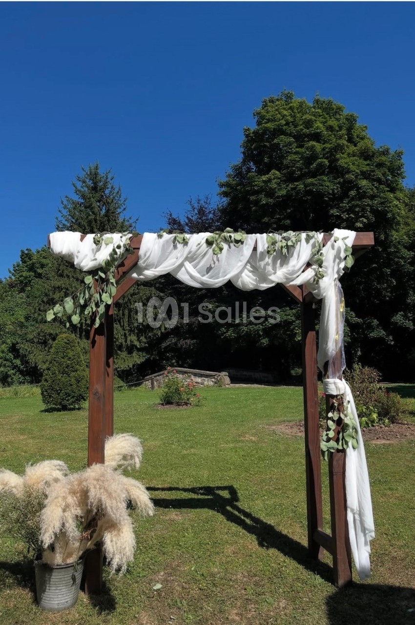 Arche en bois décorée de voilages blancs et de feuillage, installée dans un jardin sous un ciel bleu.