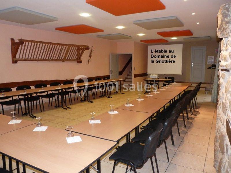 Salle de réunion aménagée en U avec tables, chaises, verres et décoration murale rustique.