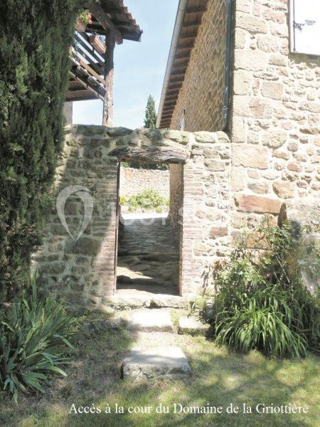 Passage en pierre menant à une cour, entouré de murs anciens, végétation et bâtiment en arrière-plan.