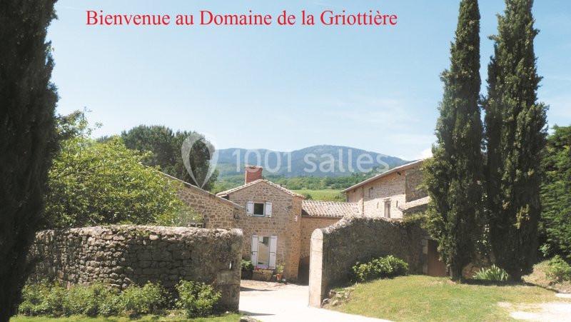 Vue d'un domaine rural avec bâtiments en pierre, arbres et montagnes en arrière-plan sous un ciel dégagé.