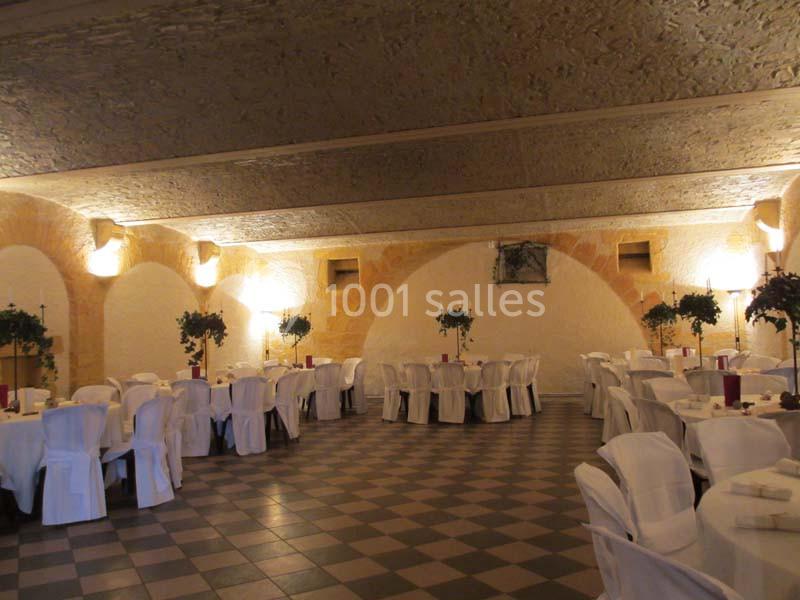 Salle de réception avec tables rondes dressées, nappes blanches et décorations végétales, éclairée par des appliques murales.