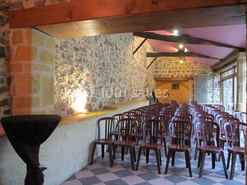 Salle en pierre avec des rangées de chaises en bois alignées, éclairée par des spots au plafond et une lumière naturelle.