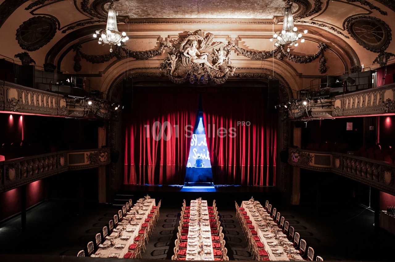 Salle de théâtre avec rideaux rouges, scène éclairée par un projecteur, tables dressées pour un événement.
