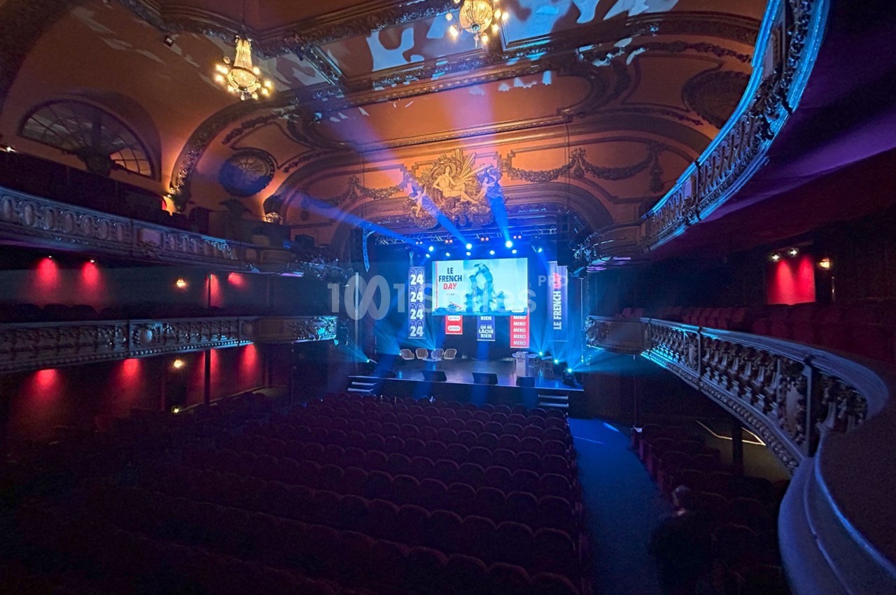 Salle de théâtre avec balcons ornés, éclairée par des lumières bleues et rouges, scène prête pour un événement.