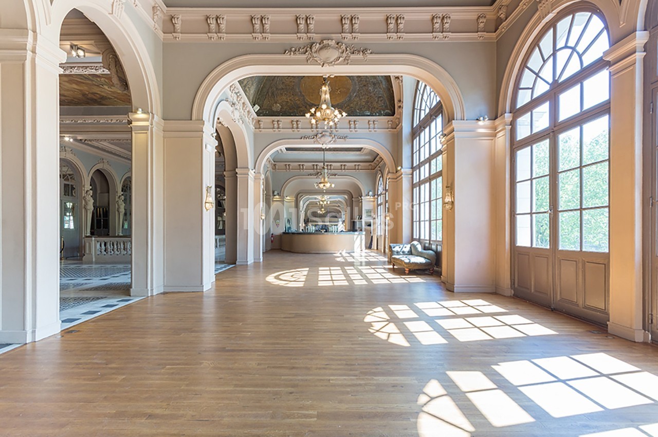 Grande salle lumineuse avec parquet, arches décoratives, lustres suspendus et larges fenêtres laissant entrer la lumière.